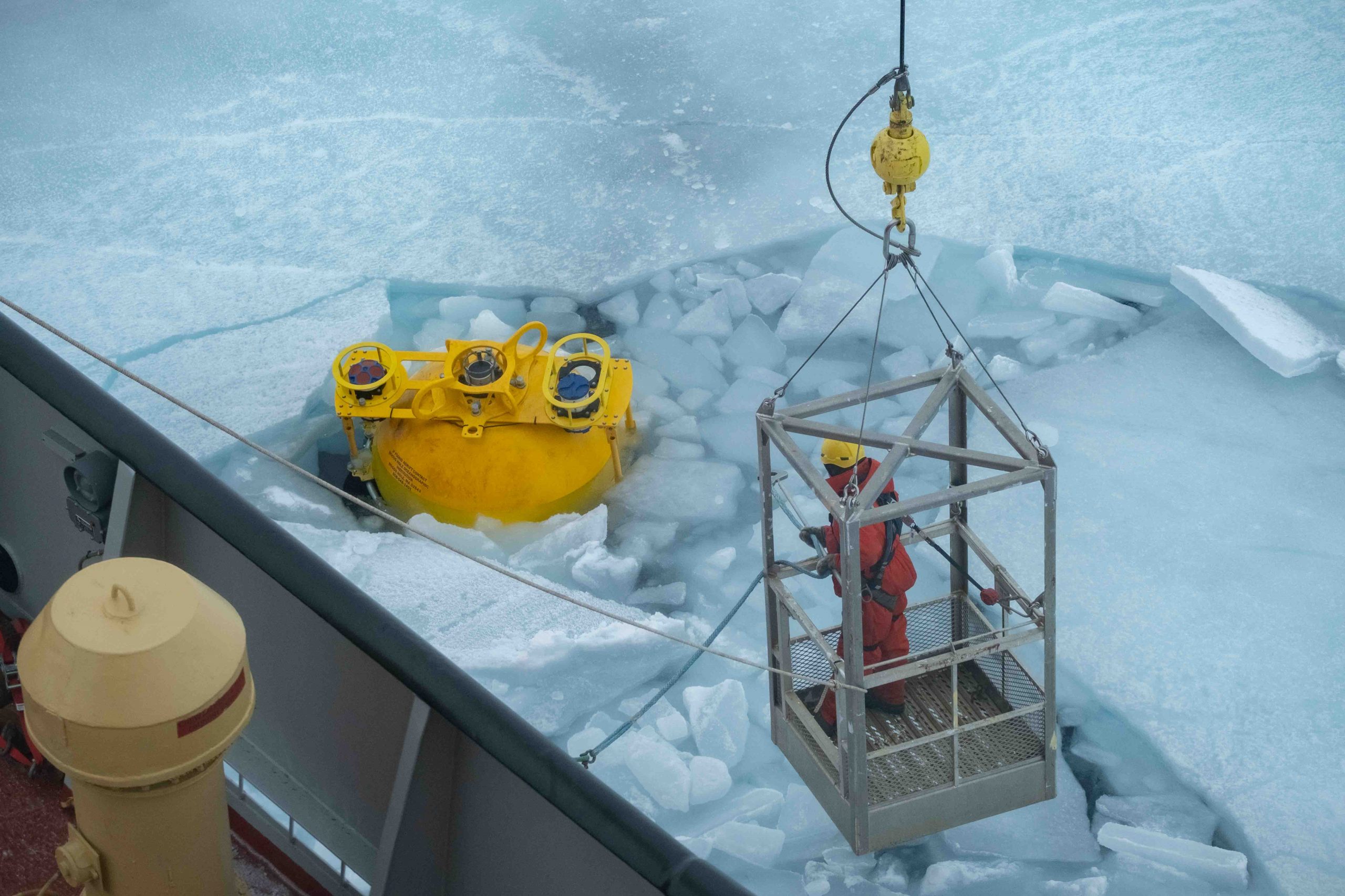 Deckhand Steve Keats in the man cage, floating over the ice to hook the surface sphere to the crane so that the mooring can begin its recovery (Photo by Elizabeth Bailey).