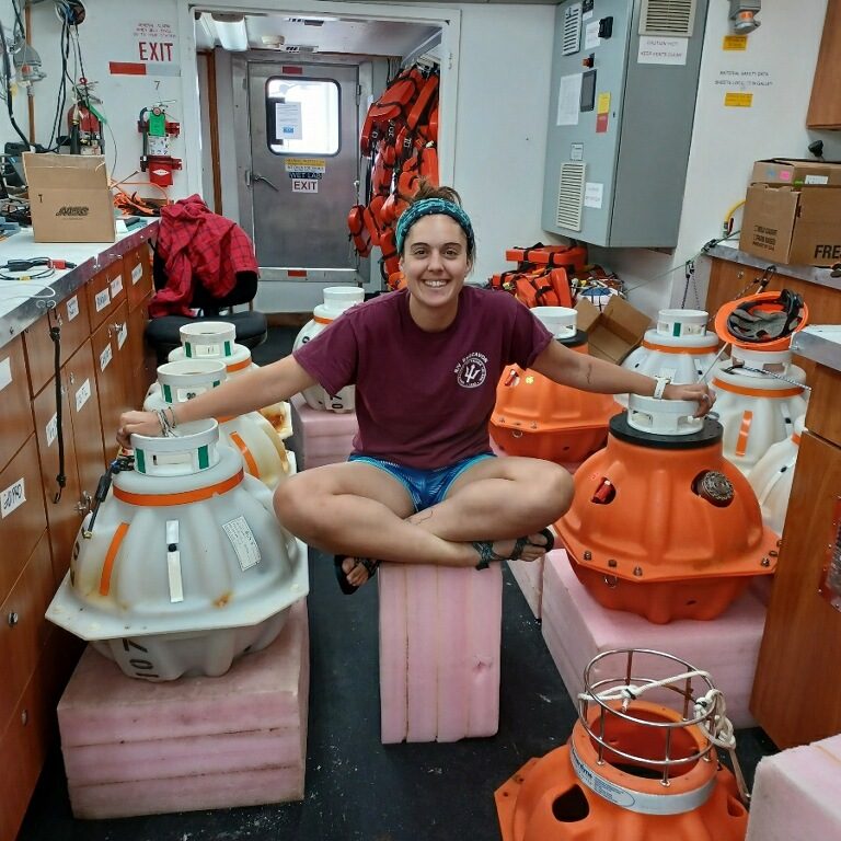 Ali sitting cross-legged in the lab of a research ship surrounded by parts of C-PIES instruments.