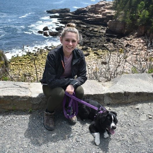 Anna and her dog sitting on a stone wall that overlooks the ocean.
