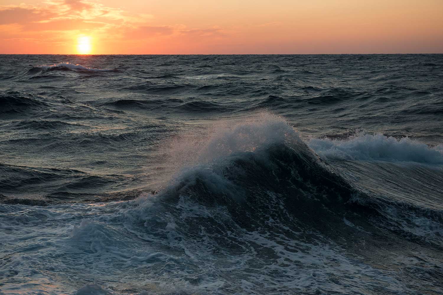 Picture shows a sunset over an open ocean with a small white-cap wave in the foreground