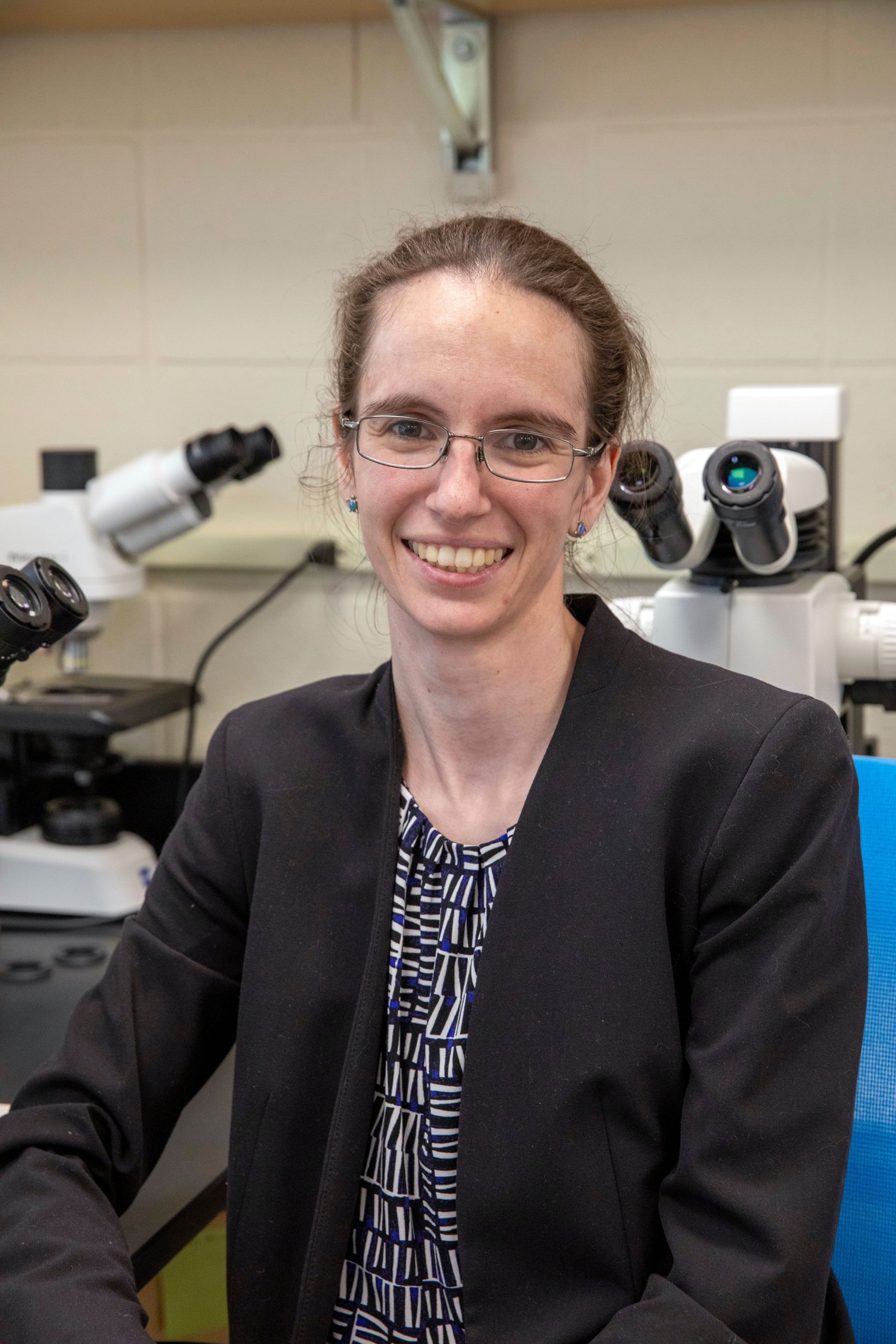 Elizabeth Sibert, a small white woman with glasses wearing a blazer, is posed sitting in the lab, in front of two microscopes, which are mostly blocked from view