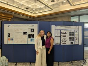 Photo is of two female undergraduate research students standing in a poster session. They are standing between two posters, posing for the camera. The woman to the left is a white woman with blonde hair wearing a white jacket and black shirt. The woman to the right is a Hispanic woman with dark hair wearing a purple top. The research posters behind them show their summer research project results