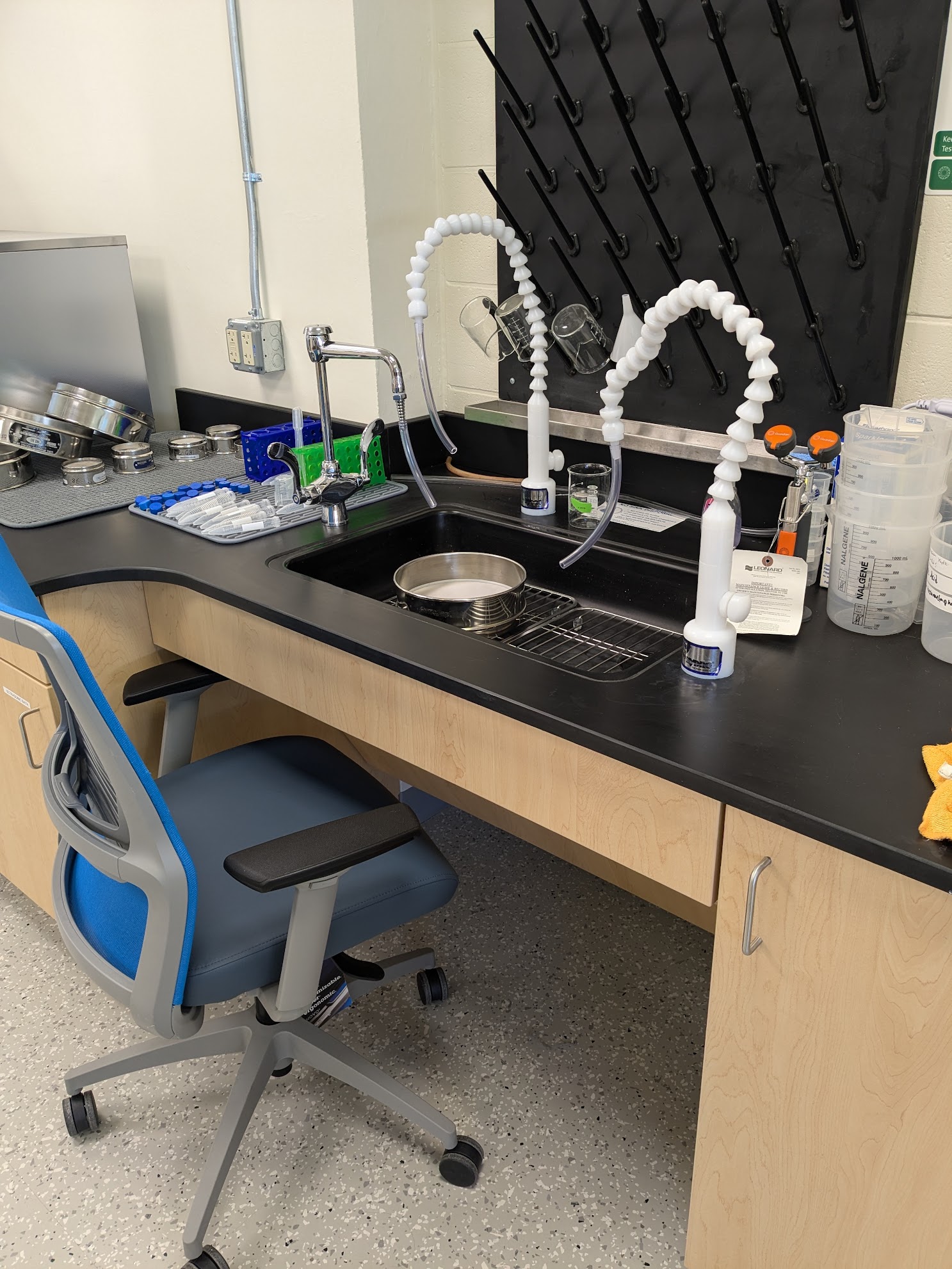 Accessible Sink is shown with chair in the knee space for scale. The sink has two white DI water faucets, as well as a tap water faucet and an eye wash. To one side of the sink is a drying station with numerous sieves and falcon tubes. To the other side of the sink are some empty beakers and DI water wash bottles. There is a beaker drying rack on the wall behind the sink, and a 8" washing sieve in the sink for scale