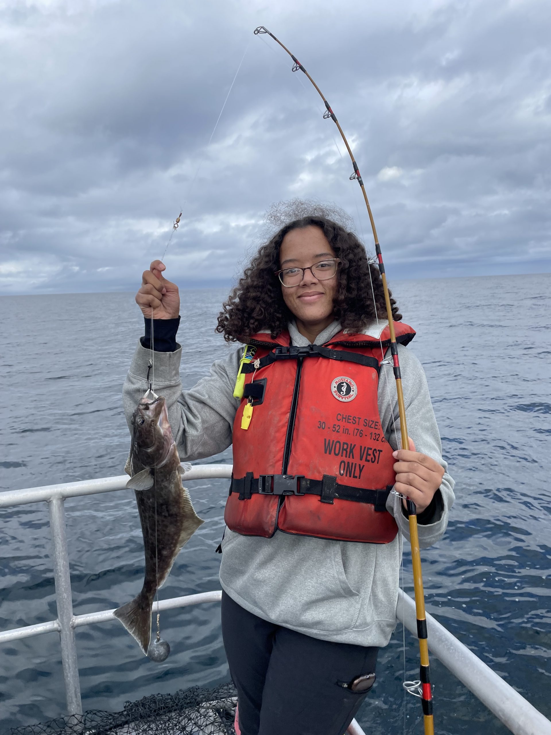 Amina Patterson, a young black woman with glasses in an orange life vest, holds a fishing rod with a freshly caught halibut on the water in Dutch Harbor Alaska.