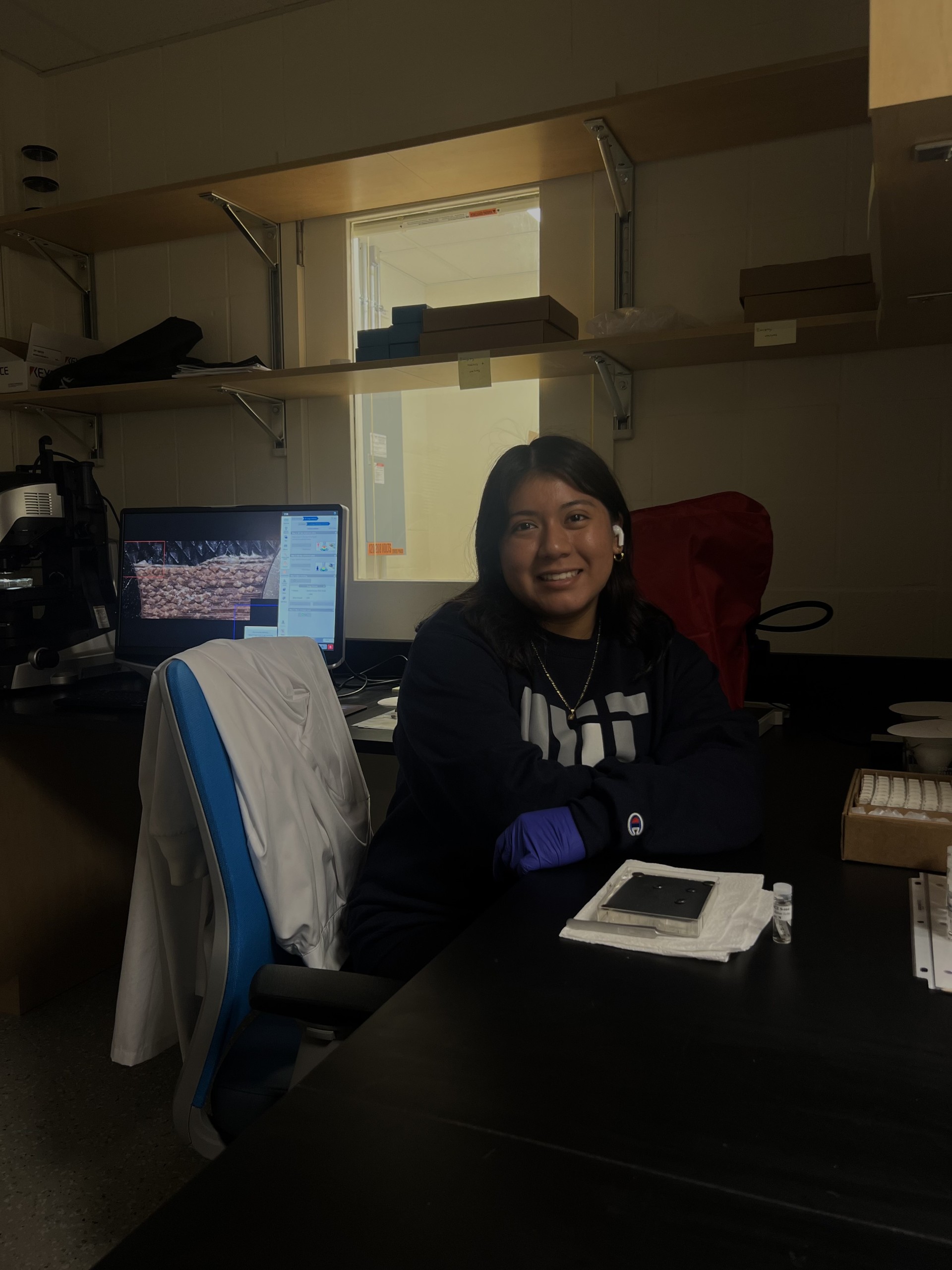 Arleth Martinez, a hispanic woman with long hair, sits in the imaging room of the paleo-FISHES Lab. She is wearing gloves and working on placing a shark skin patch on a tray to be imaged. Visible over her shoulder is the digital microscope, which is at work taking an image of a shark skin patch.