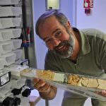 Dick Norris, a cheerful white man with a full beard, stands on a boat holding a freshly recovered deep-sea sediment core. The core contains a layer of imapct ejecta from the Cretaceous-Paleogene mass extinction event 66 million years ago that killed the dinosaurs.