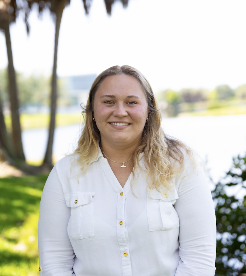 Elizabeth Pellegrini, a blonde white woman with a white collared shirt facing the viewer with a lake and palm trees in the background.