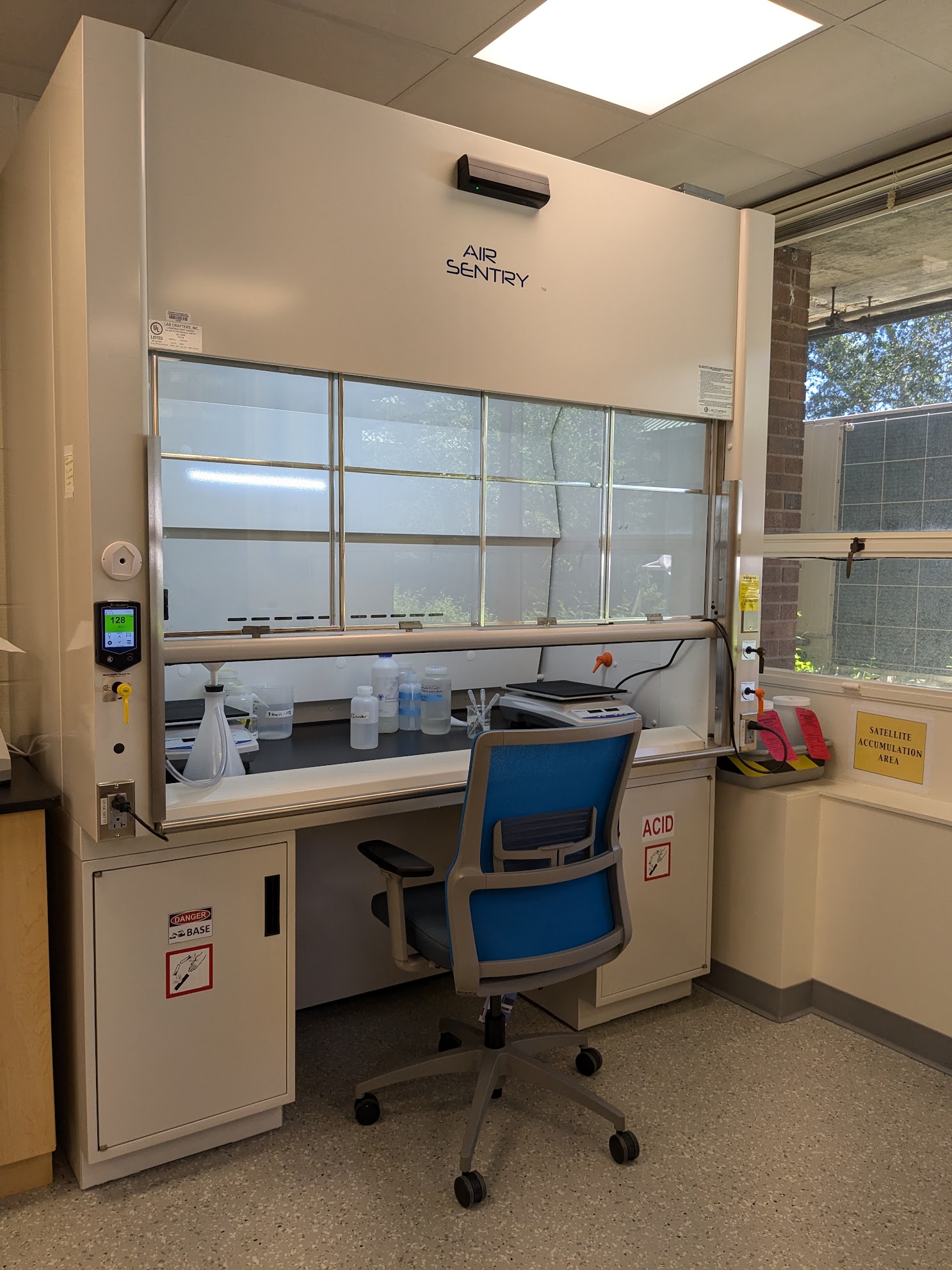 Photo shows the accessible fume hood in the Paleo-FISHES Lab space. It is located near a large window, and has a blue lab chair sitting in front of it at the workstation spot. The fume hood sash is open, and it is clear that the chair easily fits into the empty knee-space in the middle of the fume hood counter.