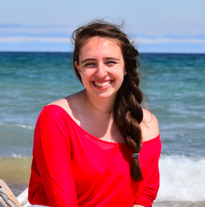 Karinne Tennenbaum, a white woman with braided brown hair, wearing a bright red shirt, posing against a blurred background of Lake Superior waves.