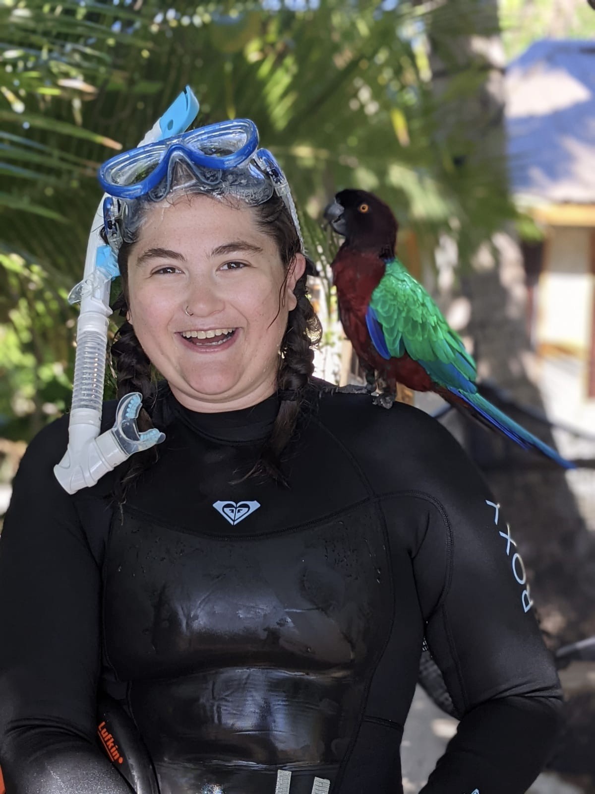 A person with wet, dark hair is wearing a black wetsuit, a snorkel and goggles. They are smiling and looking directly at the camera. They have a red and green parrot perched on their shoulder and are standing in front of a green, tropical background.