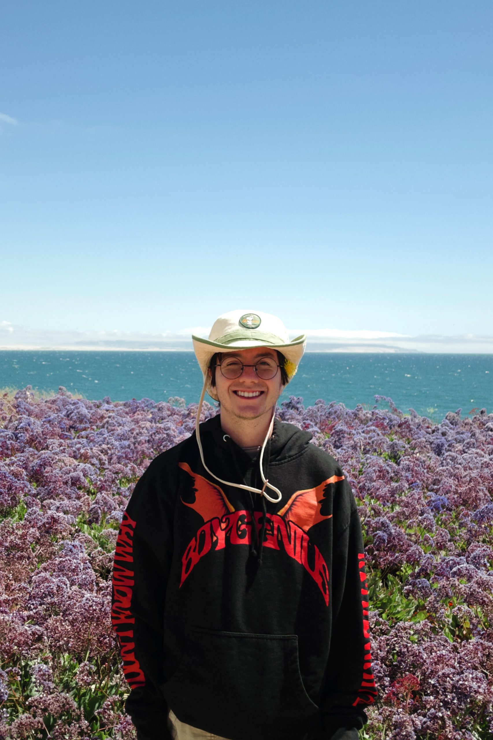 Nicholas Wallis-Mauro, a young man with glasses, wearing a hat and a black hoodie, stands in front of purple flowers, the ocean can be seen in the back.