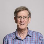 Head shot of George Lauder, an older white man with round glasses. He is smiling at the camera in front of a light gray background and wearing a blue and red plaid shirt