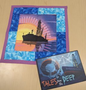 A small quilt (approximately 12 x 12 inches, with the center square showing a shilouetted image of the JOIDES Resolution, a scientific research vessel purpose built for collecting deep-sea sediments. It is pictured against an artistic version of a sunrise or sunset on the open ocean. The square central image is surrounded by blue textured fabric and a pink-purple small border. To the bottom right of the quilt is a postcard that says "Tales from the Deep: Stories of Scientific Ocean Drilling", and depects a bottom-up view (as if from underwater looking up) of the JOIDES Resolution vessel, with a life ring and a podcast microphone pointing down to the deep sea.