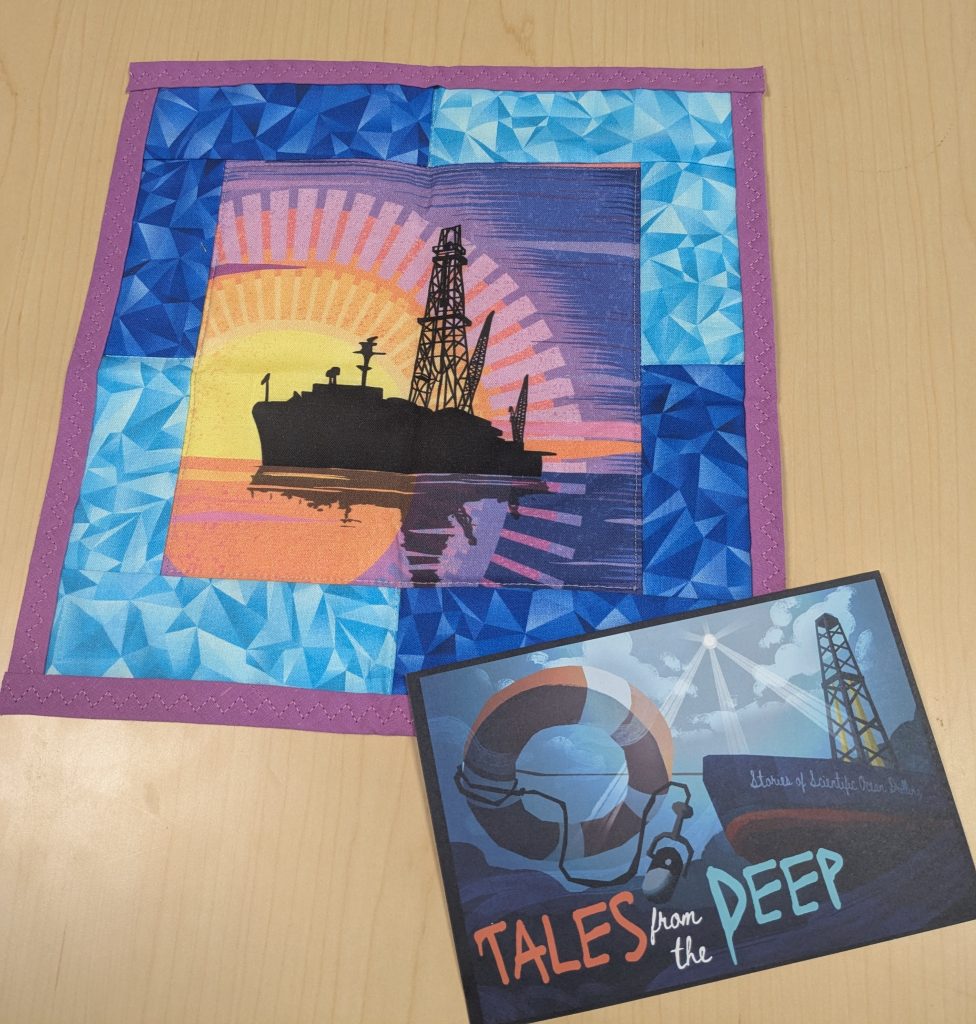 A small quilt (approximately 12 x 12 inches, with the center square showing a shilouetted image of the JOIDES Resolution, a scientific research vessel purpose built for collecting deep-sea sediments. It is pictured against an artistic version of a sunrise or sunset on the open ocean. The square central image is surrounded by blue textured fabric and a pink-purple small border. To the bottom right of the quilt is a postcard that says "Tales from the Deep: Stories of Scientific Ocean Drilling", and depects a bottom-up view (as if from underwater looking up) of the JOIDES Resolution vessel, with a life ring and a podcast microphone pointing down to the deep sea.
