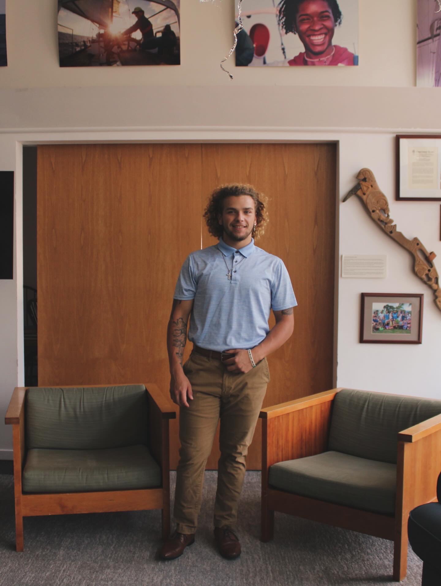 Alex Brown, a average heighted mixed gentlemen that is clean shaven, standing in between two chairs in a open windowed room wearing brown shoes, brown cacky pants, a light blue collared shirt