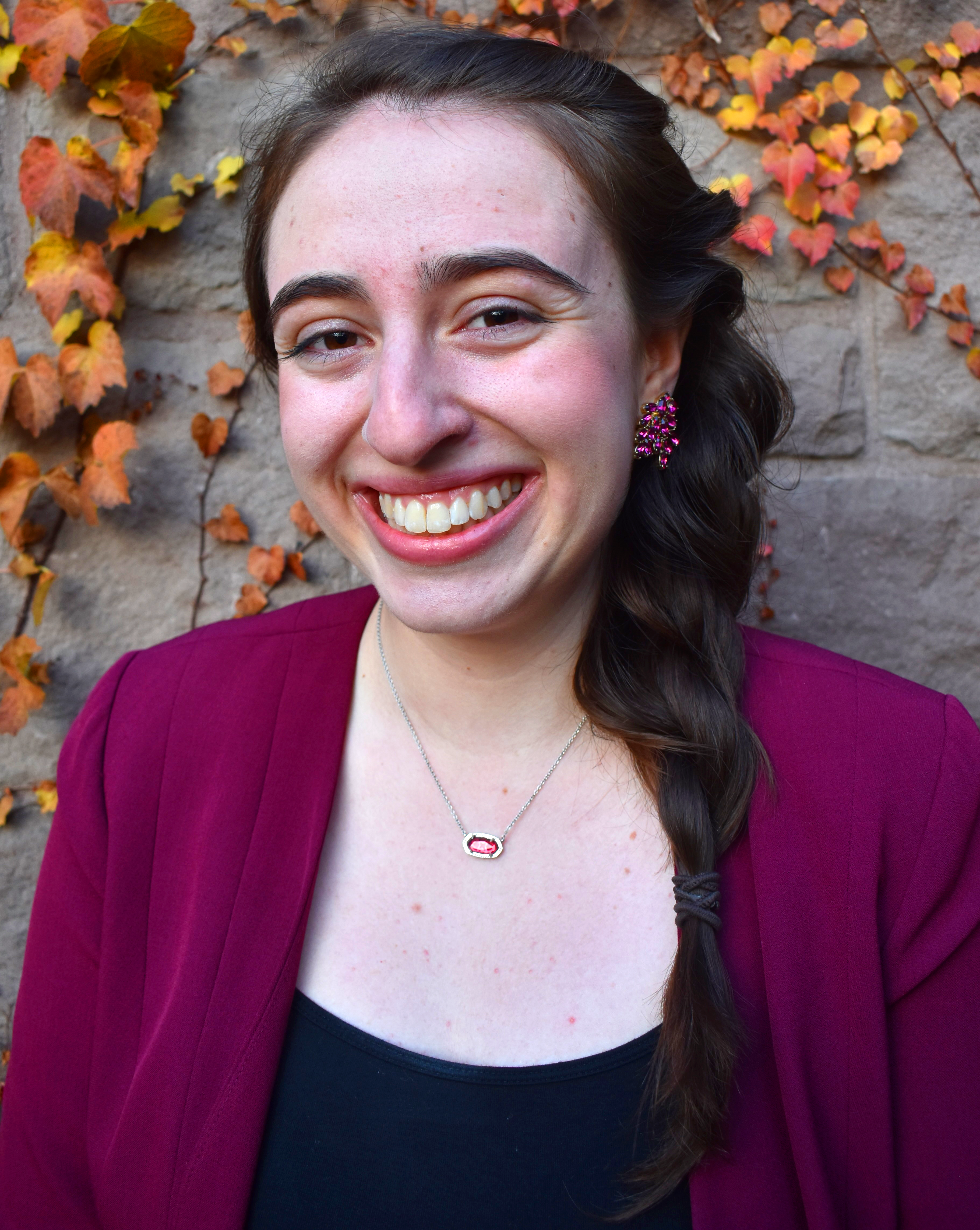 Headshot of Karinne Tennenbaum, a white woman with long braided brown hair. She is standing in front of a gray sone wall
