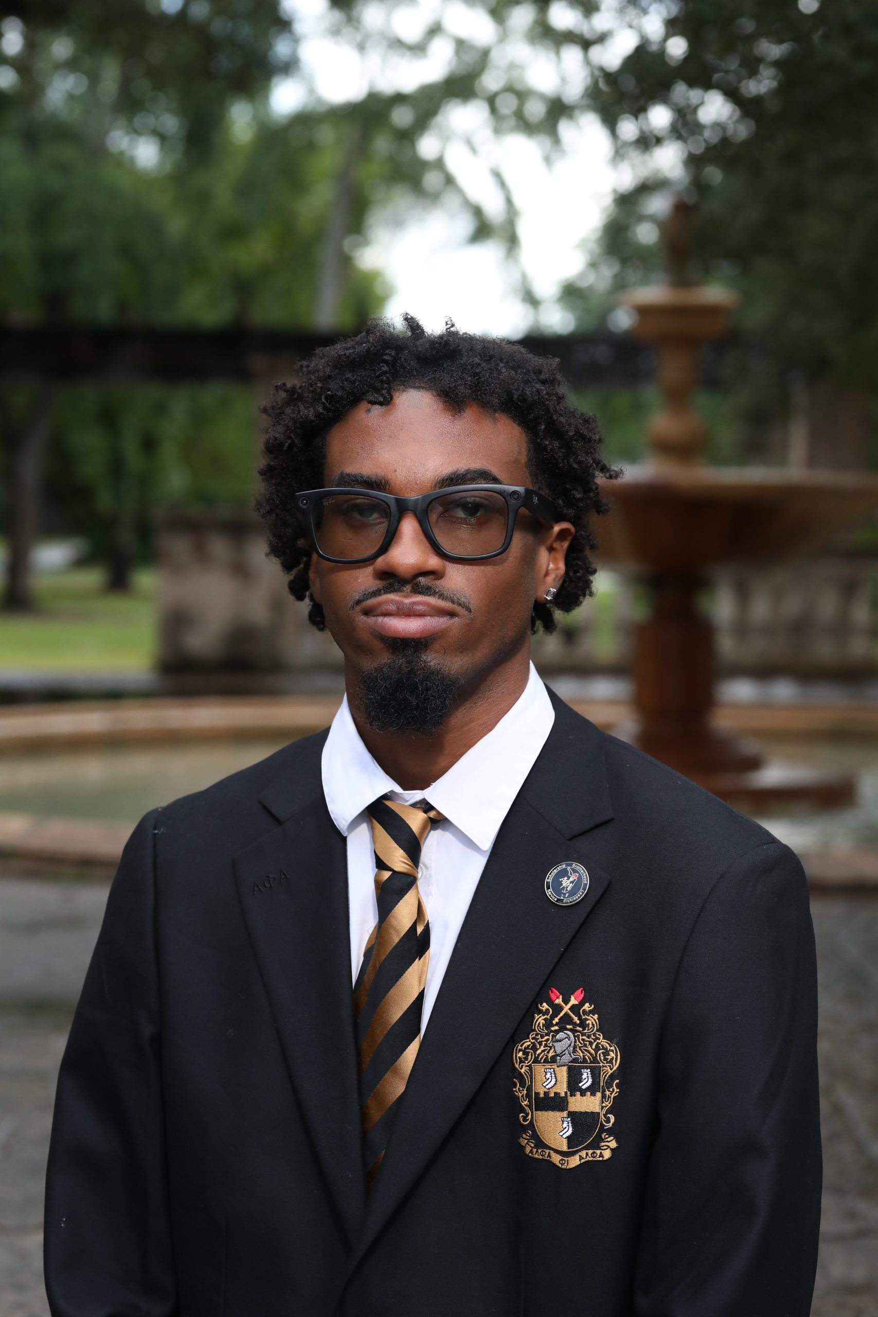 Akil stands outside in front of a blurred background. He is a tall black man, wearing a blazer and tie.