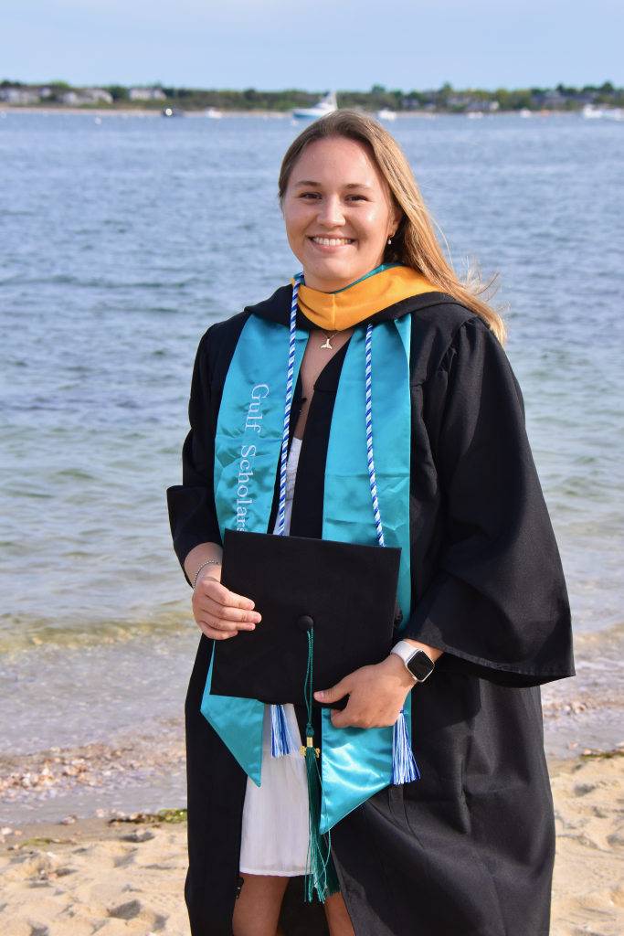 Elizabeth Pellegrini is a white woman with long blonde hair. She is standing on a beach in full graduation regalia, and holding a diploma.