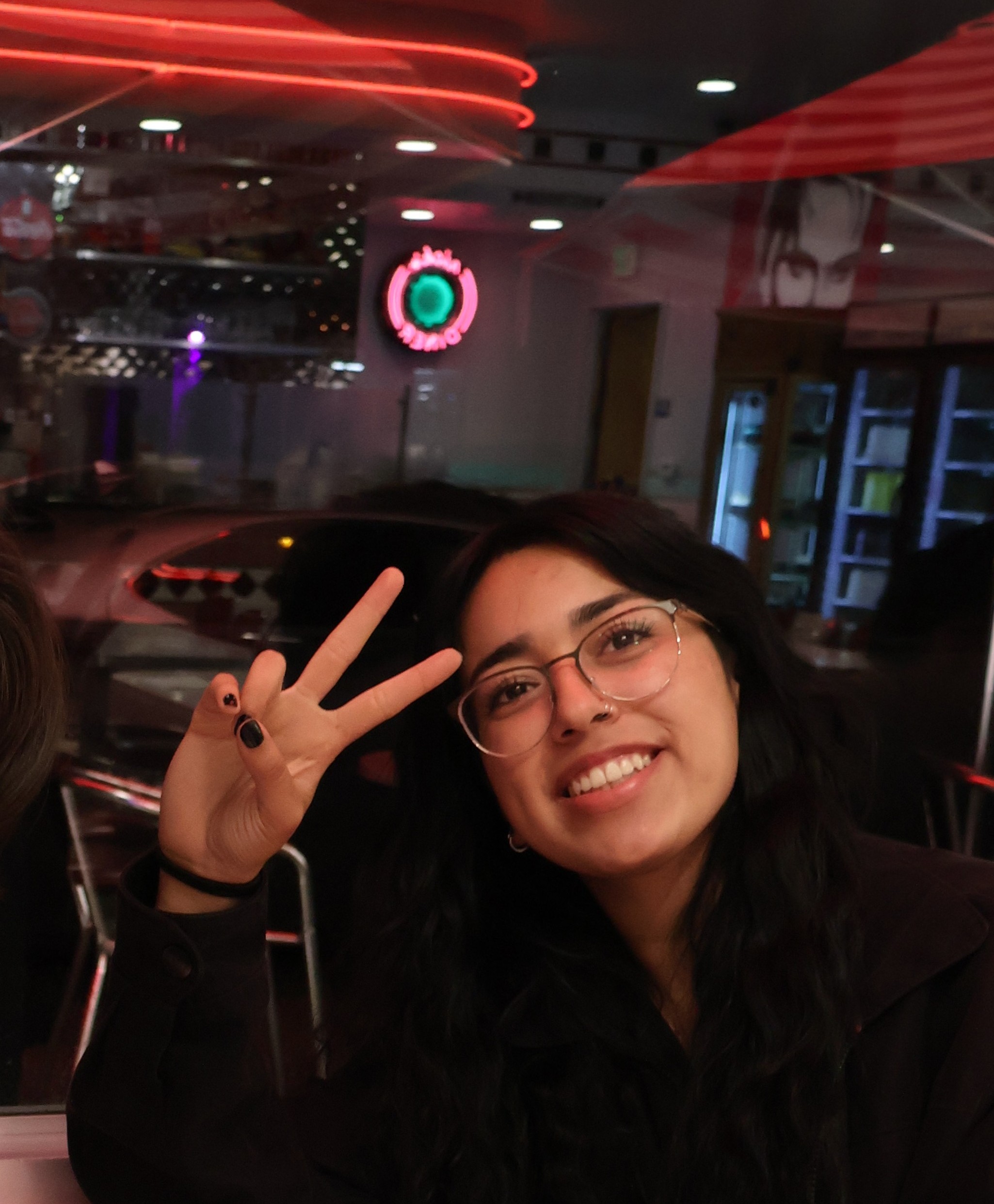 Keila Alejos is a young woman with long dark brown hair and glasses. She is pictured in a dark room with fluorescent lights and is opsing for the camera