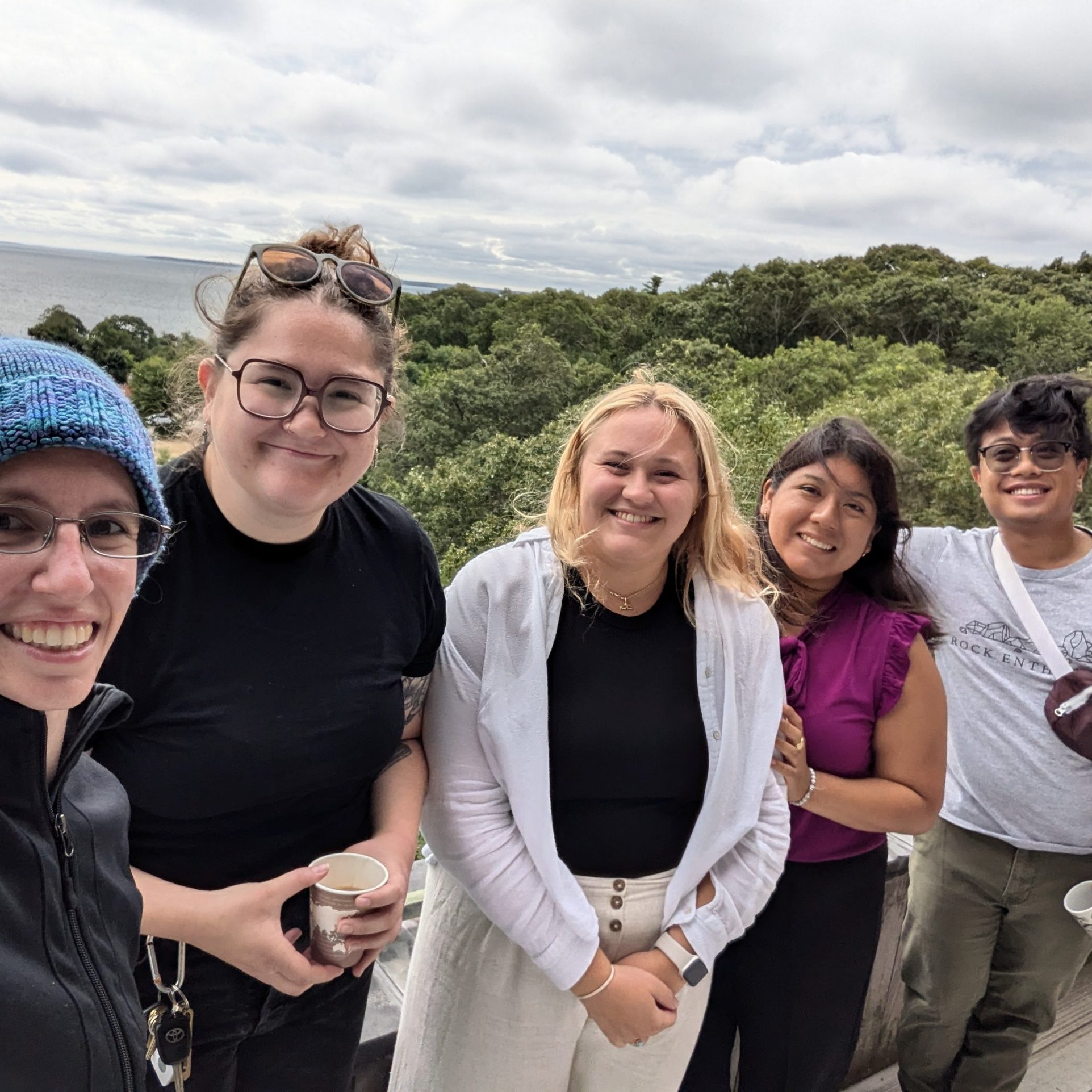 Lab selfie with 5 people taken from the balcony of Clark Labs 5th floor. There are 5 people lined up along the wall of the balcony, with trees and the ocean below