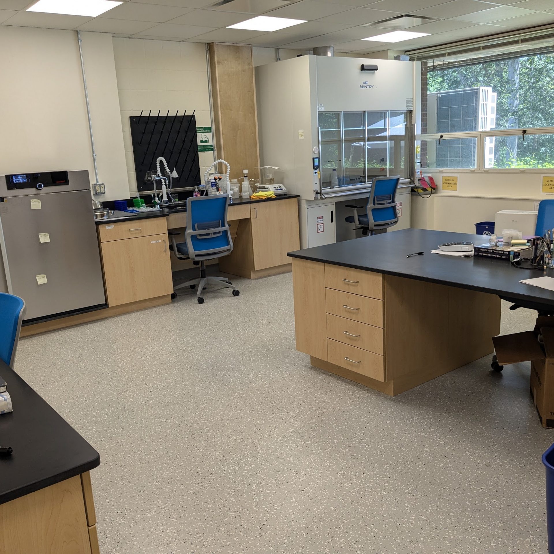 View of the lab from the side door, showing the wetlab wall (fume hood, sink, oven, standign height counter) to the left, and the microscope and seated-height desk working stations to the right