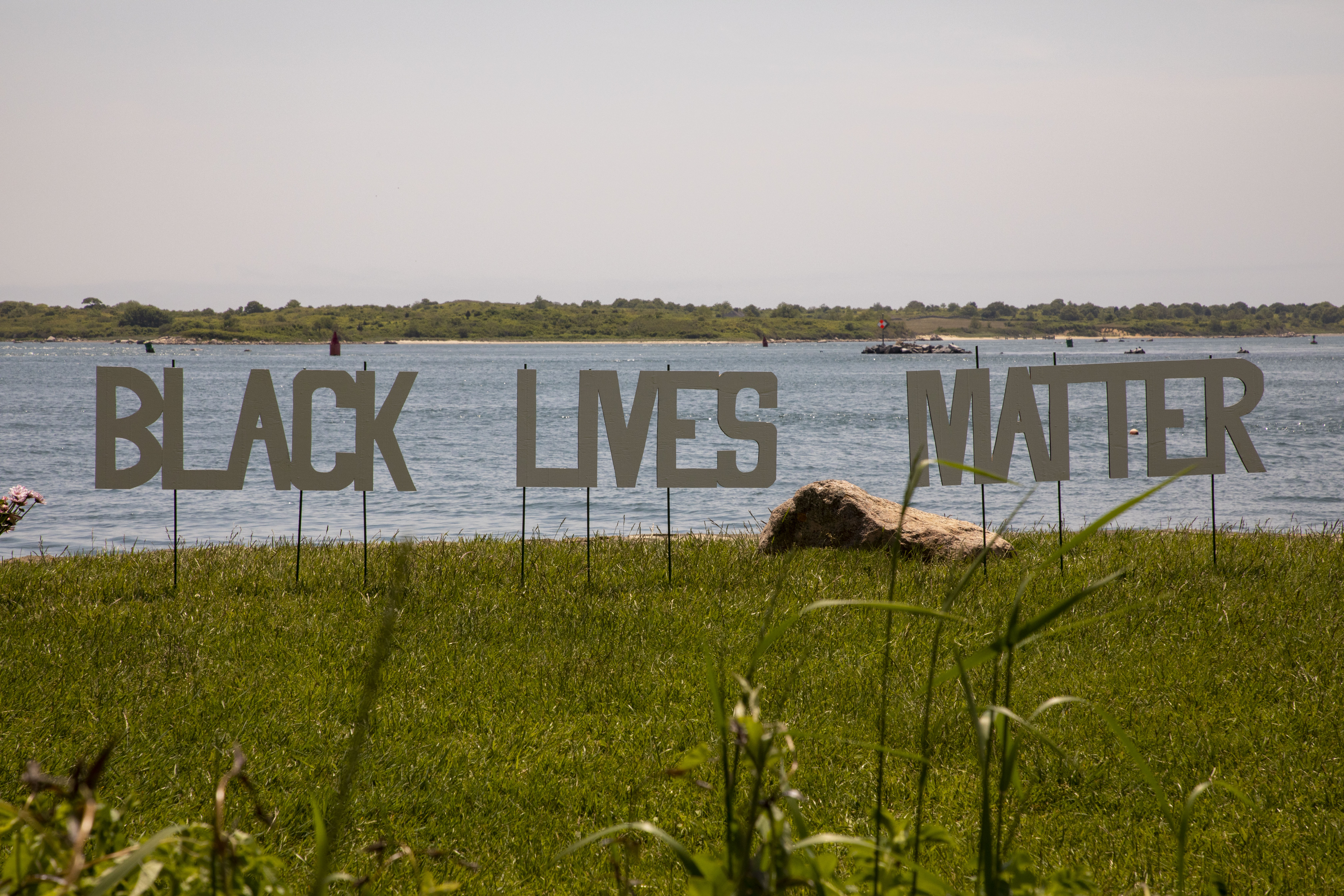 Photography of block letter cutouts on stakes planted in the grass spelling out "Black Lives Matter". The picture is composed in thirds, low third is green grass, middle third is the words against a background of blue water, and the top third is a milky overcast sky.