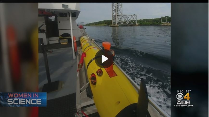 Photo of a yellow tube shaped robot in a cradle on the deck of a boat going under a bridge on the canal.